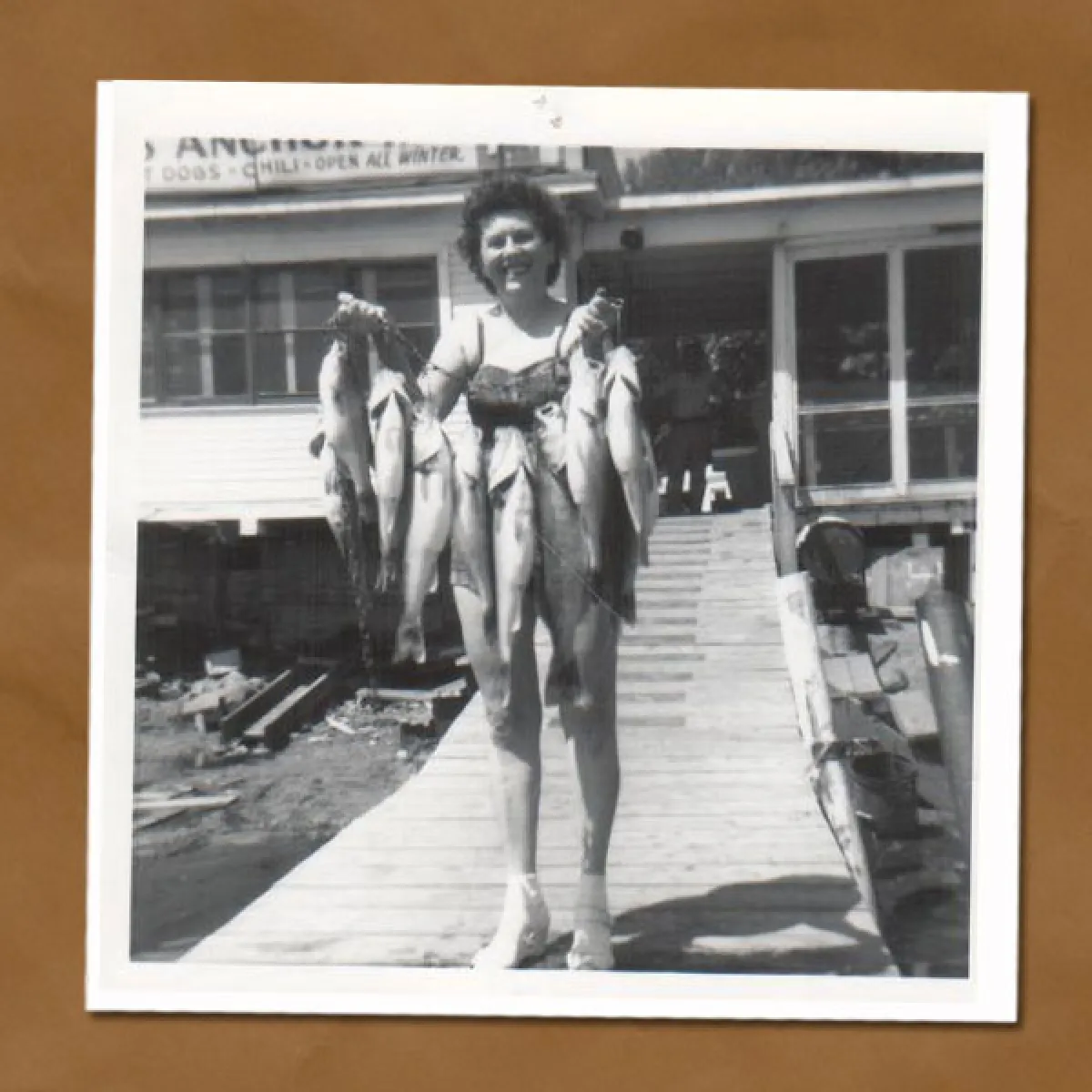Black and white photo of woman standing on pier holding up string of fish she just caught